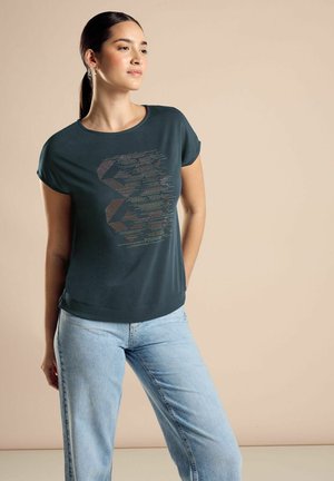 T-shirt verde scuro a maniche corte con un design geometrico testurizzato in colori contrastanti sulla parte anteriore, abbinato a jeans blu chiaro.