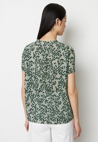 Kurzärmlige Bluse aus hellgrünem Stoff mit dunkelgrünem Punktenmuster, mit rundem Ausschnitt und subtilen Falten im Rücken.