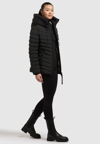 Schwarze Daunenjacke mit Kapuze, vertikaler Steppstruktur, zwei vorderen Reißverschlusstaschen und glänzender Innenauskleidung. Getragen mit schwarzen Leggings und Stiefeln.
