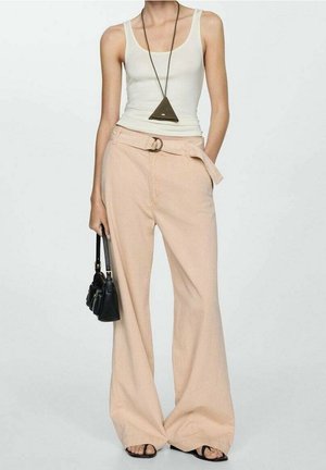 Osoba w białym topie na ramiączkach, beżowych szerokich spodniach z paskiem, czarnych sandałach, z naszyjnikiem z trójkątnym wisiorkiem oraz trzymająca małą czarną torebkę.