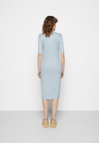 Vestido azul claro, justo, até ao joelho, com mangas curtas. Textura lisa, design minimalista e tecido suave. Calçado são tamancos bege.