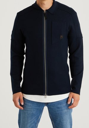 Chaqueta azul marino con cremallera y cuello, que presenta un bolsillo en el pecho, textura acanalada y una capa interior blanca en contraste visible en el dobladillo.