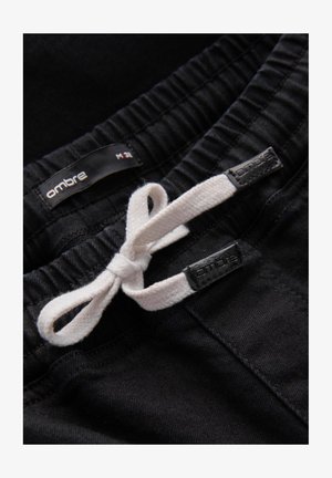 Zwarte broek met een getextureerde stof, voorzien van een elastische tailleband en een witte trekkoord. Labels met merknaam "ombre" zichtbaar.