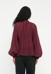 Bluză burgundy cu o croială lejeră, mâneci tip balon și închidere cu nasturi la spate. Material fin cu volane de-a lungul decolteului.