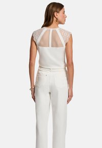 Top sans manches blanc avec des accents en dentelle et en mesh aux épaules et dans le dos. Associé à un pantalon blanc taille haute. Texture lisse et design ajusté.