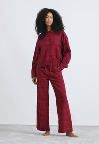 Rød rutete pysjamas-sett som har en knapp-up skjorte med krage og løstsittende bukser med elastisk liv. Stoffet ser mykt ut.