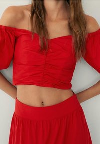 Haut court rouge à épaules dénudées avec une texture froncée. Manches courtes bouffantes et ourlet droit. Associé à une jupe rouge assortie à la taille.