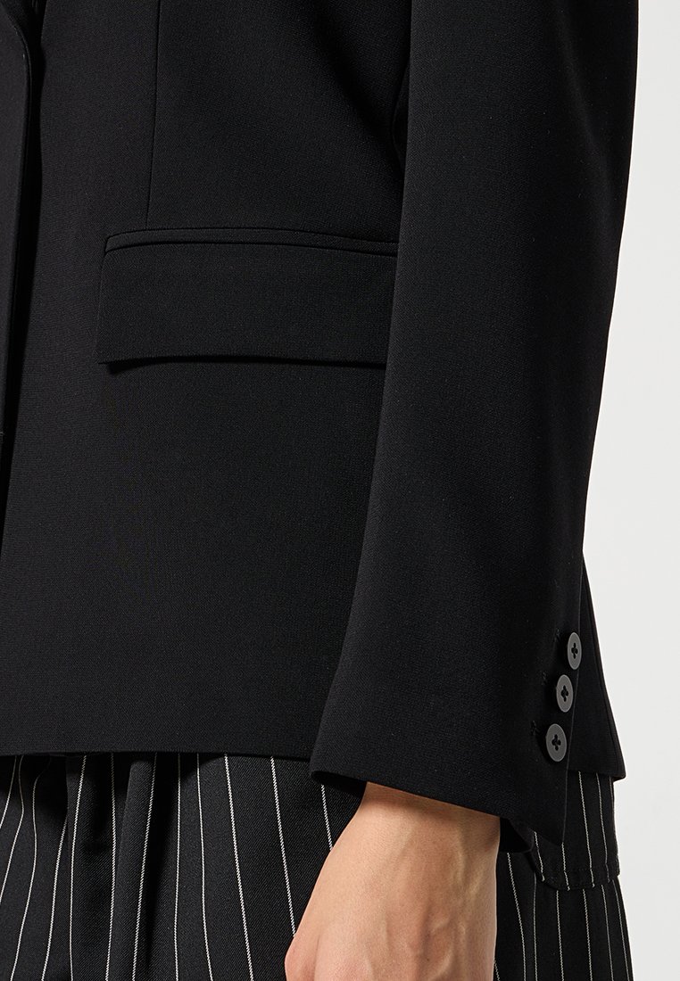 Gros plan sur la manche d'un blazer noir avec trois boutons et une poche à rabat, porté avec un pantalon noir à rayures, montrant le poignet et la main.