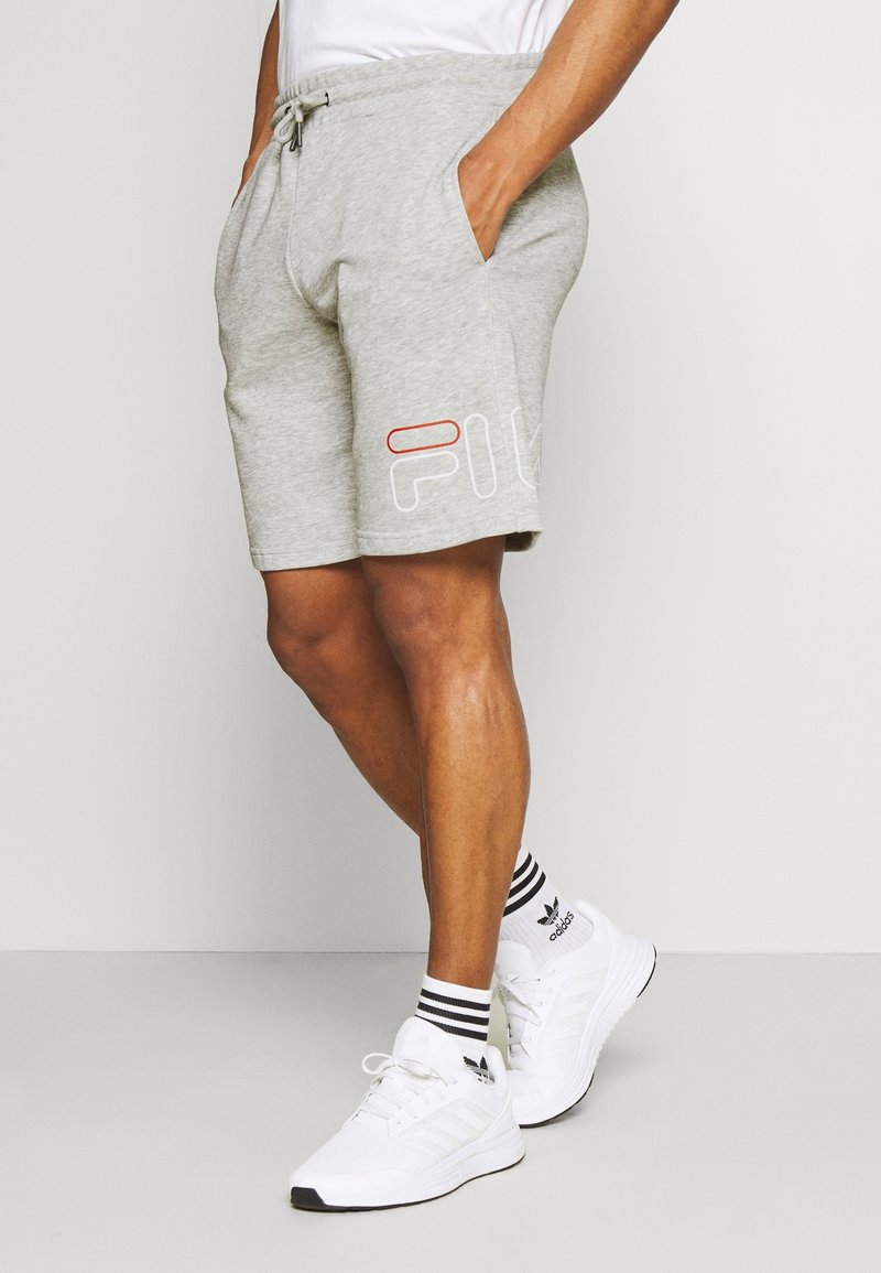 Shorts gris en mélange de coton avec une ceinture élastique, des poches latérales et un détail de logo blanc et rouge sur la jambe gauche. Associés à des baskets blanches.
