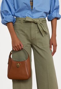 Brown leather handbag with a curved shape, gold hardware, and a top handle, paired with olive green high-waisted pants and a blue blouse.