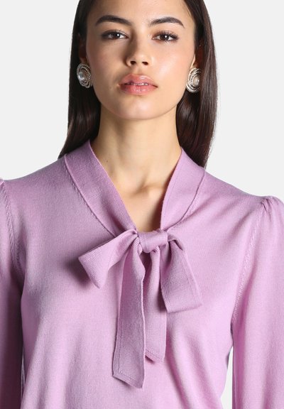 Jeune femme aux cheveux lisses et foncés porte un haut violet clair avec un col noué en nœud et de grandes boucles d'oreilles en perles.