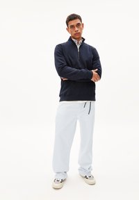 Marineblauwe rits sweatshirt met geribbelde manchetten en zoom, gedragen over een wit shirt, gecombineerd met lichtblauwe losse broek en beige sneakers.