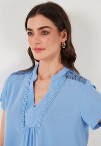 Blusa blu con maniche corte, scollatura a V, dettagli in pizzo decorativi sulle spalle e un design frontale arricciato. Tessuto morbido.