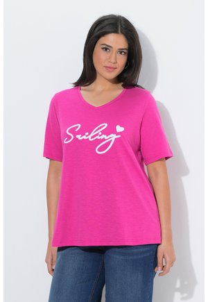 Kvinde iført en knaldpink T-shirt med V-hals med teksten "Smiling" og et hjertegrafik, stående mod en ensfarvet hvid baggrund, iført blå jeans.