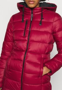 Mujer vistiendo una chaqueta acolchada roja con capucha, cremallera negra y bolsillo vertical en el pecho, con su mano derecha en el bolsillo de la chaqueta.