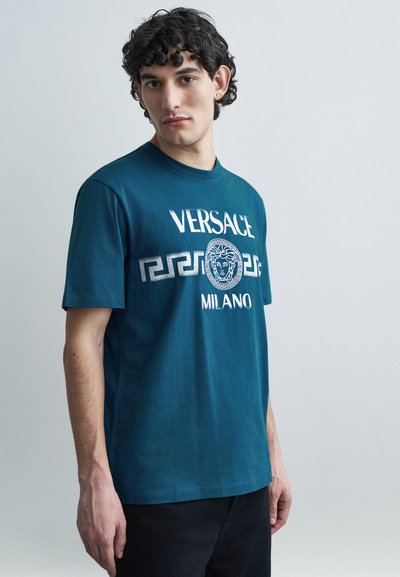 T-shirt en coton bleu sarcelle avec grand texte blanc "VERSACE", un logo graphique et un motif clé grec. Manches courtes et col rond.