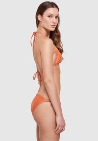 Oranje bikini set met een striksluiting achter. De top heeft driehoekige cups, terwijl de onderkant laag zit met minimale bedekking. Gladde textuur.