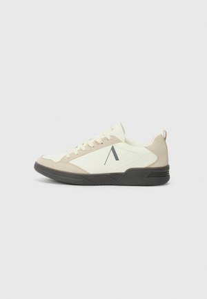 Sneaker med overdel i hvit lær og beige semsket skinn. Har en svart gummisåle, rund tå og et minimalistisk logodesign.