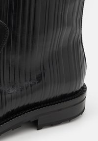 Botte en cuir noir avec une texture côtelée, dotée d'une semelle en caoutchouc robuste, de coutures visibles et d'un design à bout arrondi.