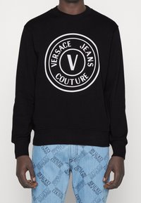 Schwarzer Pullover mit großem weißem Kreislogo, das den Text "VERSACE JEANS COUTURE" und ein auffälliges "V" in der Mitte zeigt.