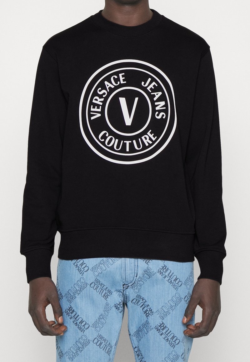 Schwarzer Pullover mit großem weißem Kreislogo, das den Text "VERSACE JEANS COUTURE" und ein auffälliges "V" in der Mitte zeigt.