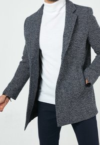 Marineblauw-witte wollen jas met een patroon, voorzien van een reverskraag, open voorkant en zijzakken, gedragen over een witte coltrui.