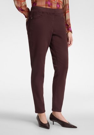 Donna con pantaloni slim fit marrone scuro, scarpe marroni con tacco basso e punta affilata, e camicia a quadri a maniche lunghe, con le mani nelle tasche.