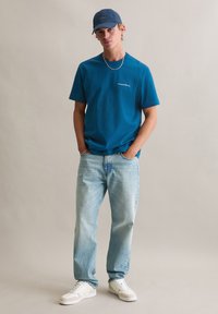 Niebieski bawełniany T-shirt z małym białym logo, zestawiony z jasnoniebieskimi jeansami i białymi trampkami. Model nosi dopasowaną niebieską czapkę.