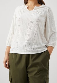 Biała bluzka z ażurowymi detalami, okrągłym dekoltem i rękawami 3/4, z pionowymi wzorami; połączona z luźnymi spodniami w oliwkowym kolorze.