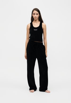 Jeune femme portant un haut noir sans manches et un pantalon noir large, debout pieds nus sur un fond clair uni.