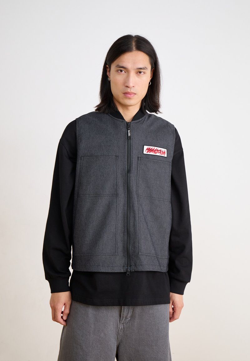 Gray fabric vest with a front zipper, two large front pockets, and a red patch. Worn over a black long-sleeve shirt, featuring a relaxed fit.