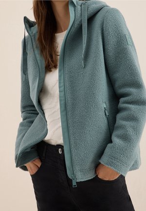 Femme portant un sweat à capuche zippé en polaire bleu sarcelle clair, les mains dans les poches d’un pantalon noir et un pull texturé blanc en dessous.