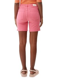 Persona vestida con pantalones cortos de mezclilla rosa y sandalias beige, de pie con la espalda hacia la cámara contra un fondo blanco.