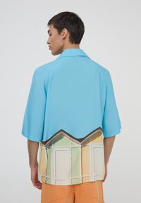 Camicia a maniche corte in tessuto azzurro chiaro con un design colorato di capanne sulla spiaggia lungo l'orlo, con un profilo morbido e un colletto classico.
