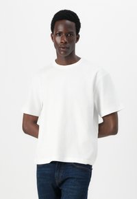 White short-sleeve t-shirt in a loose fit, made of cotton with a crew neck. Worn with blue jeans, with a plain and smooth texture.