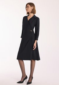 Czarna sukienka typu wrap z długimi rękawami, kołnierzykiem i detalem guzika z boku. Posiada dopasowany krój i spódnicę do kolan, wykonaną z gładkiego materiału.