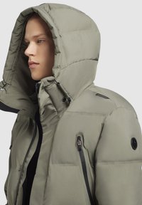 Jachetă tip puffer de culoare verde oliv deschis, având o glugă mare, buzunare cu fermoar și o textură netedă, ușor lucioasă. Accente negre vizibile.