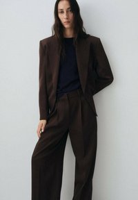 Costume marron ajusté avec un pantalon large et un blazer structuré. Porté sur un haut noir ajusté, mettant en valeur une silhouette élancée et un design moderne.