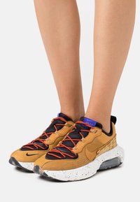 Nike sneakers med en överdel i tan mocka, svart mesh, orange snören och en vit fläckad sula med en synlig luftkammare.