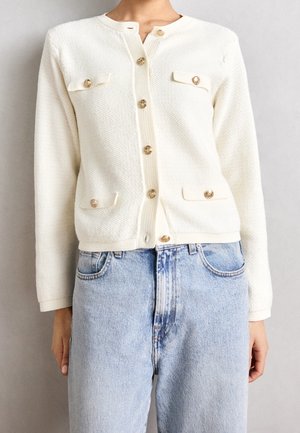 Cardigan en maille blanche texturée avec boutons dorés et rabats de poche, porté avec un jean taille haute bleu clair.