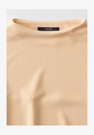 Haut couleur pêche clair fabriqué en tissu lisse avec un col montant et des manches courtes. Présente une étiquette noire portant la mention "un jour."