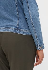 Person som bär en blå jeansjacka och olivgröna byxor, med jackans ärm knäppt vid handleden och handen avslappnad vid sidan.