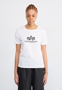 Valkoinen lyhythihainen puuvillapaita, jossa on musta graafinen logo "ALPHA INDUSTRIES" ja vaakasuorat viivat sen alla. Yksinkertainen, pyöreä kaula-aukko.