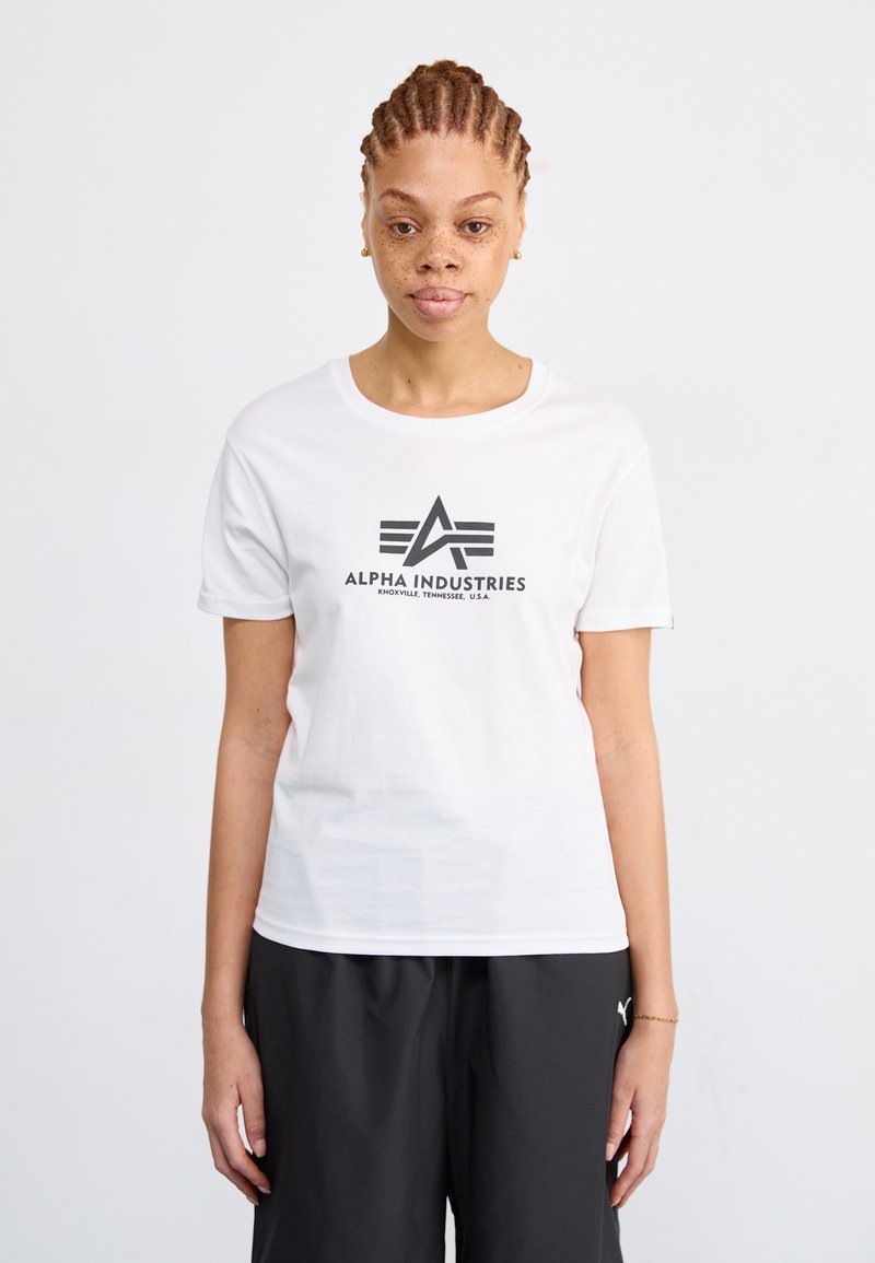 Valkoinen lyhythihainen puuvillapaita, jossa on musta graafinen logo "ALPHA INDUSTRIES" ja vaakasuorat viivat sen alla. Yksinkertainen, pyöreä kaula-aukko.