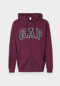 Bordeaux-kleurige zip-up hoodie met een voorzak, voorzien van een groot "GAP" logo in wit en marineblauw over de borst. Glad stof, verstelbare capuchon.