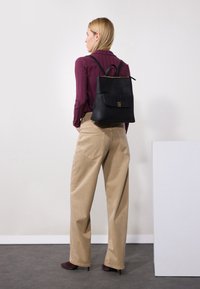 Black leather backpack with a top flap and gold hardware. Tapered, beige trousers and dark shoes complete the outfit. Vertical striped shirt in maroon.