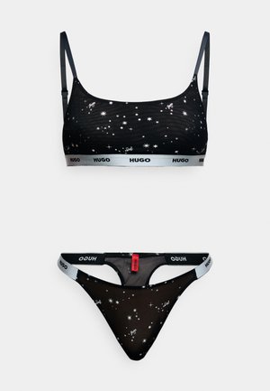 Schwarzes Lingerie-Set bestehend aus einem Bralette und einem Tanga mit Sternmustern, transparentem Stoff und elastischen Bändern mit "HUGO"-Logo in weißen und roten Akzenten.