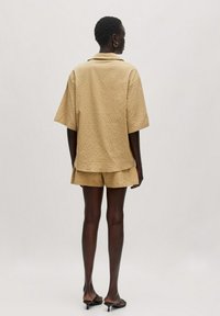 Beige strukturiertes Kurzarmhemd mit Kragen, kombiniert mit passenden Shorts. Das Model trägt schwarze Absatzsandalen und hat kurze Haare.