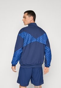 Giacca atletica blu navy con un sovrapposto geometrico blu sulle spalle e sulla schiena, polsini a costine e colletto alto, abbinata a pantaloni corti blu.