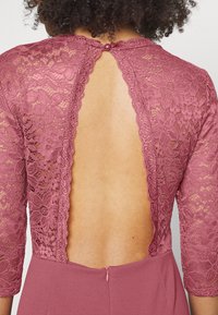 Vestido de renda em rosa poeirento com costas transparentes e mangas de três quartos. Apresenta um fecho de botão no pescoço e um fecho oculto na cintura.